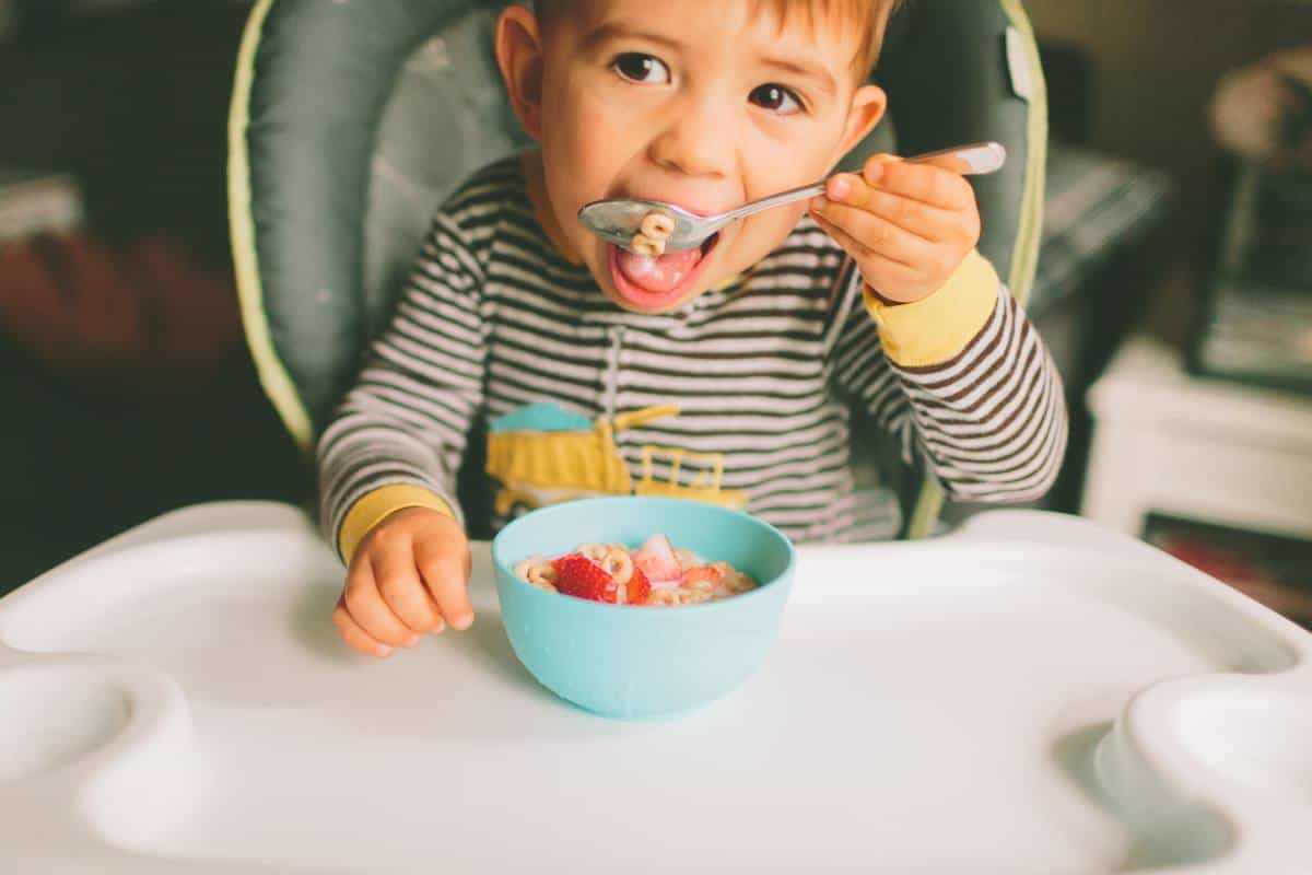 A toddler in a high chair enjoying a bowl of cheerios with sliced strawberries.