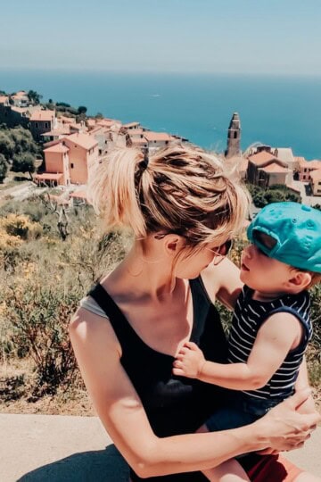 Mama holding toddler in an Italian town with a view of the Mediterranean in the background.